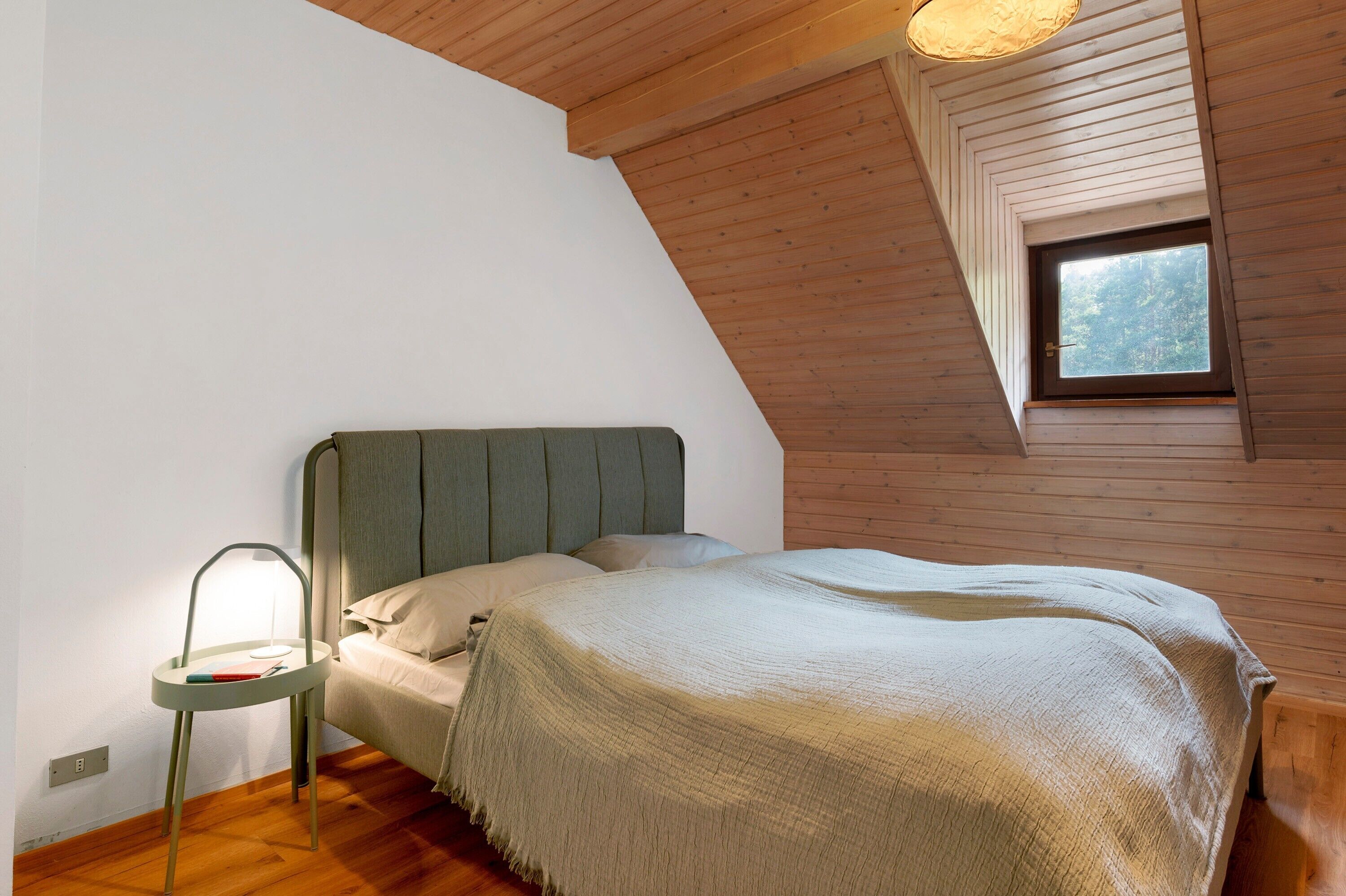 A bedroom with a bed, a bedside table with a lamp, wooden walls, and a window.