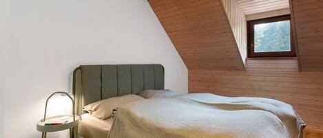 A bedroom with a bed, a bedside table with a lamp, wooden walls, and a window.