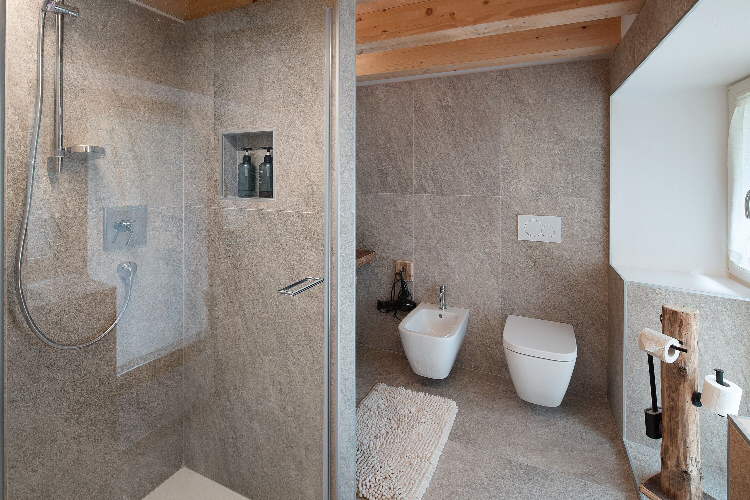 A modern bathroom with a shower, bidet, toilet, and a wooden toilet paper holder.