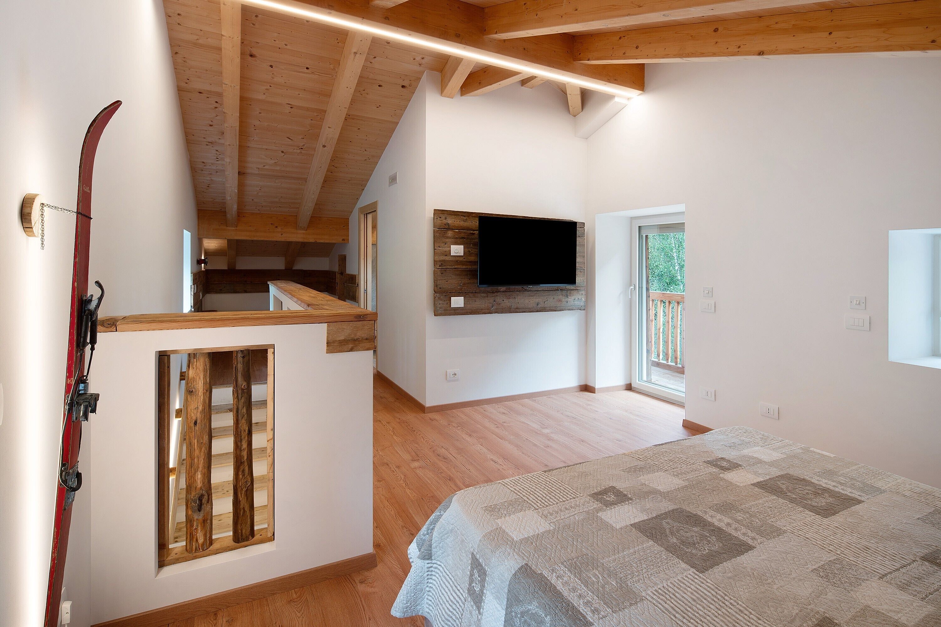 A bedroom with a bed, a mounted TV, wooden beams, a window, and a ski rack.