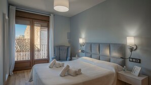 Un dormitorio con cama, mesita de noche, luces fijadas a la pared y vistas de edificios a través de una ventana.