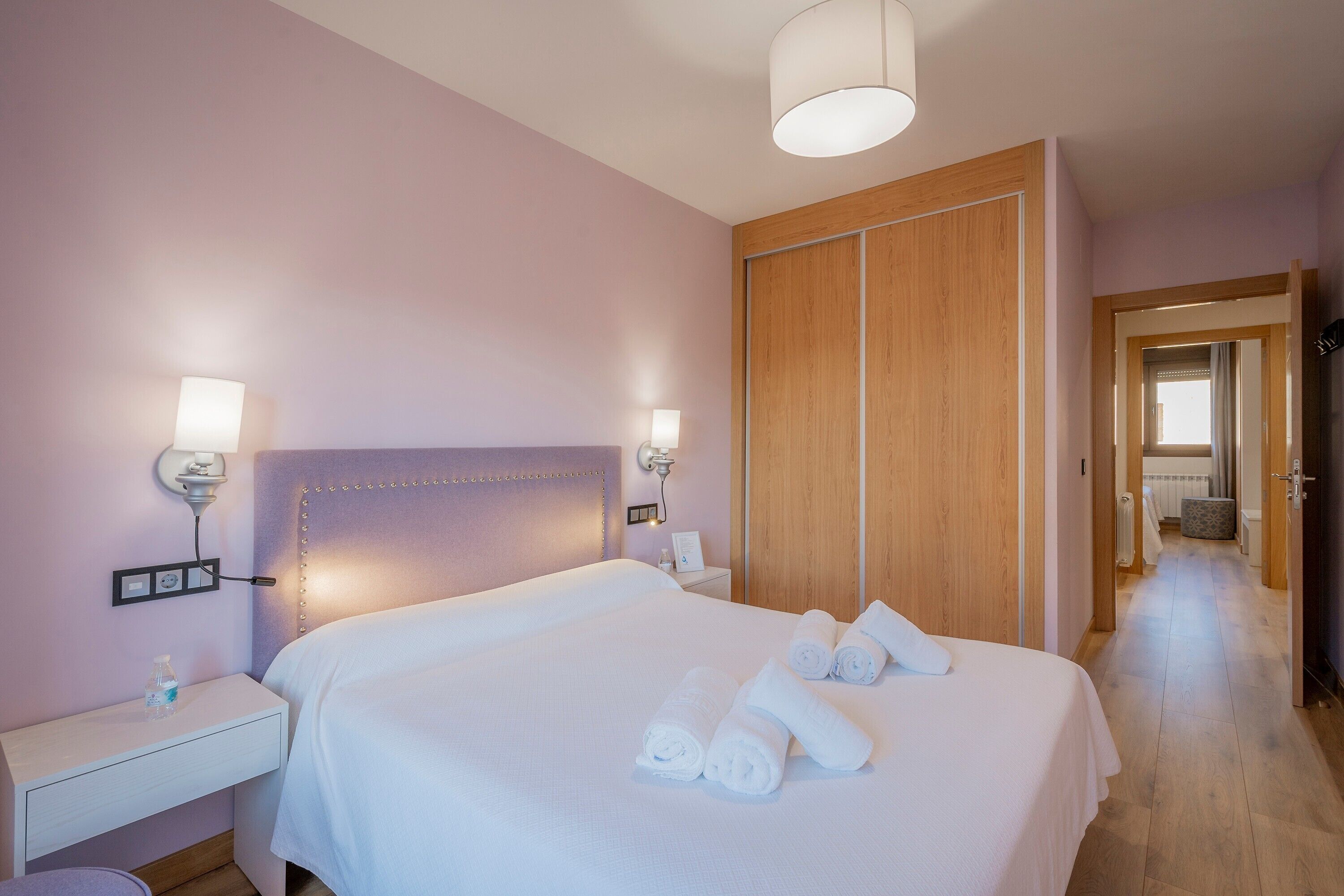 Una camera d'albergo con un letto, comodini, lampade a parete, un armadio e un corridoio che conduce a un'altra stanza.