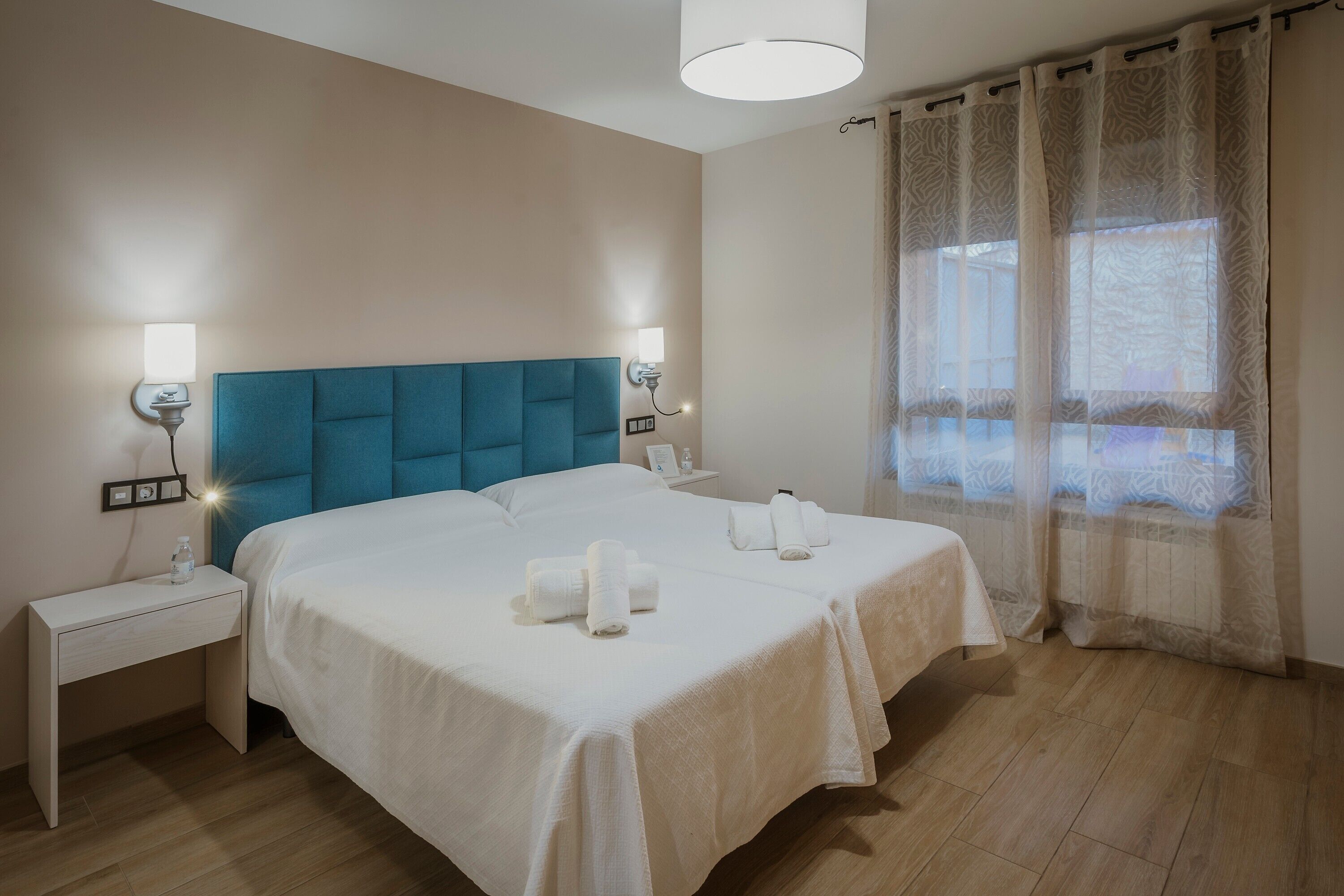 Une chambre d’hôtel avec deux lits, une table de chevet, une lampe, une fenêtre avec des rideaux et un luminaire au plafond.