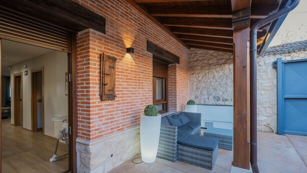 Fachada de ladrillo con techo de madera, una puerta y una zona de estar con cojín y una planta pequeña.