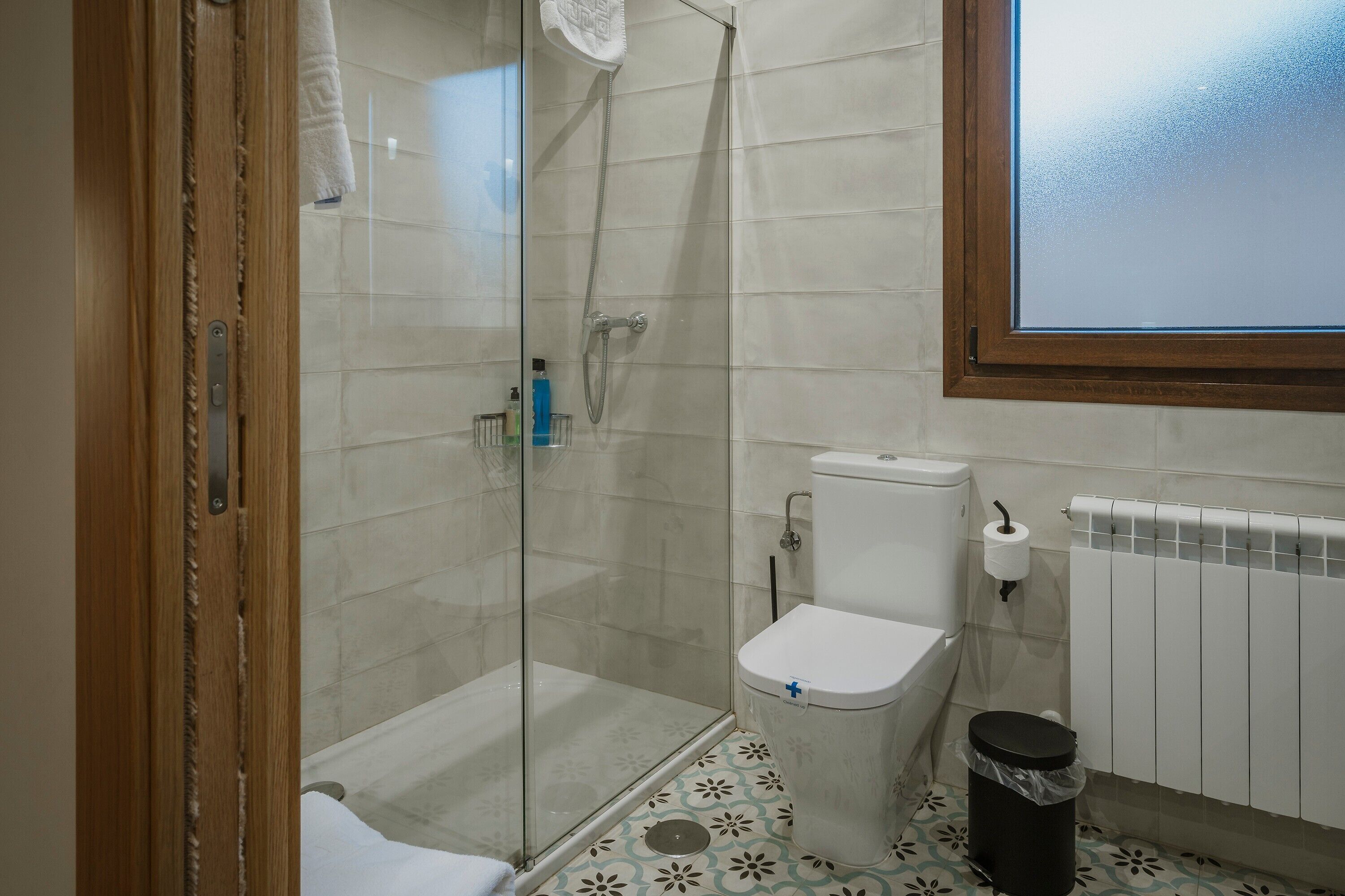 A bathroom with a glass-enclosed shower, a toilet, a radiator, and a trash bin.
