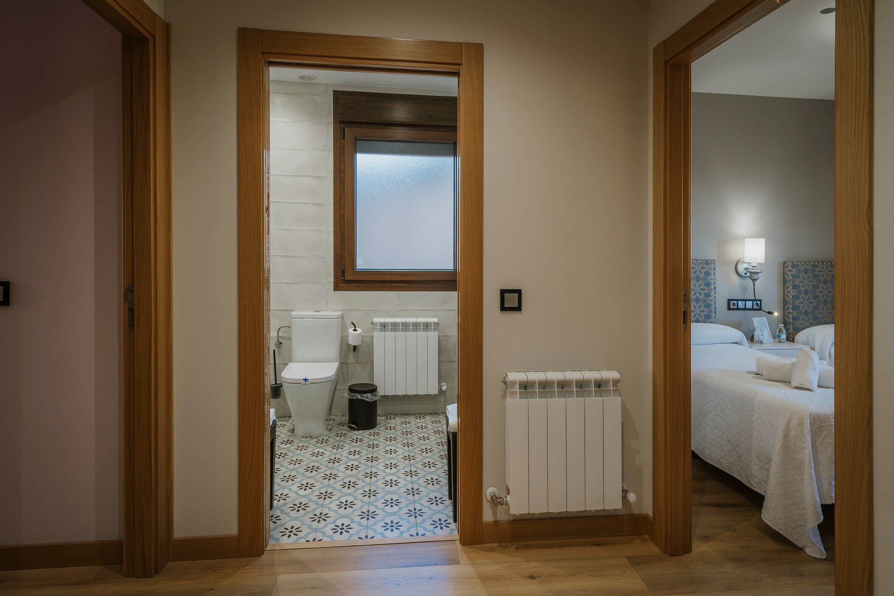 A room with a wooden door, a window, a radiator, a bed, and a bathroom with a toilet and a sink.