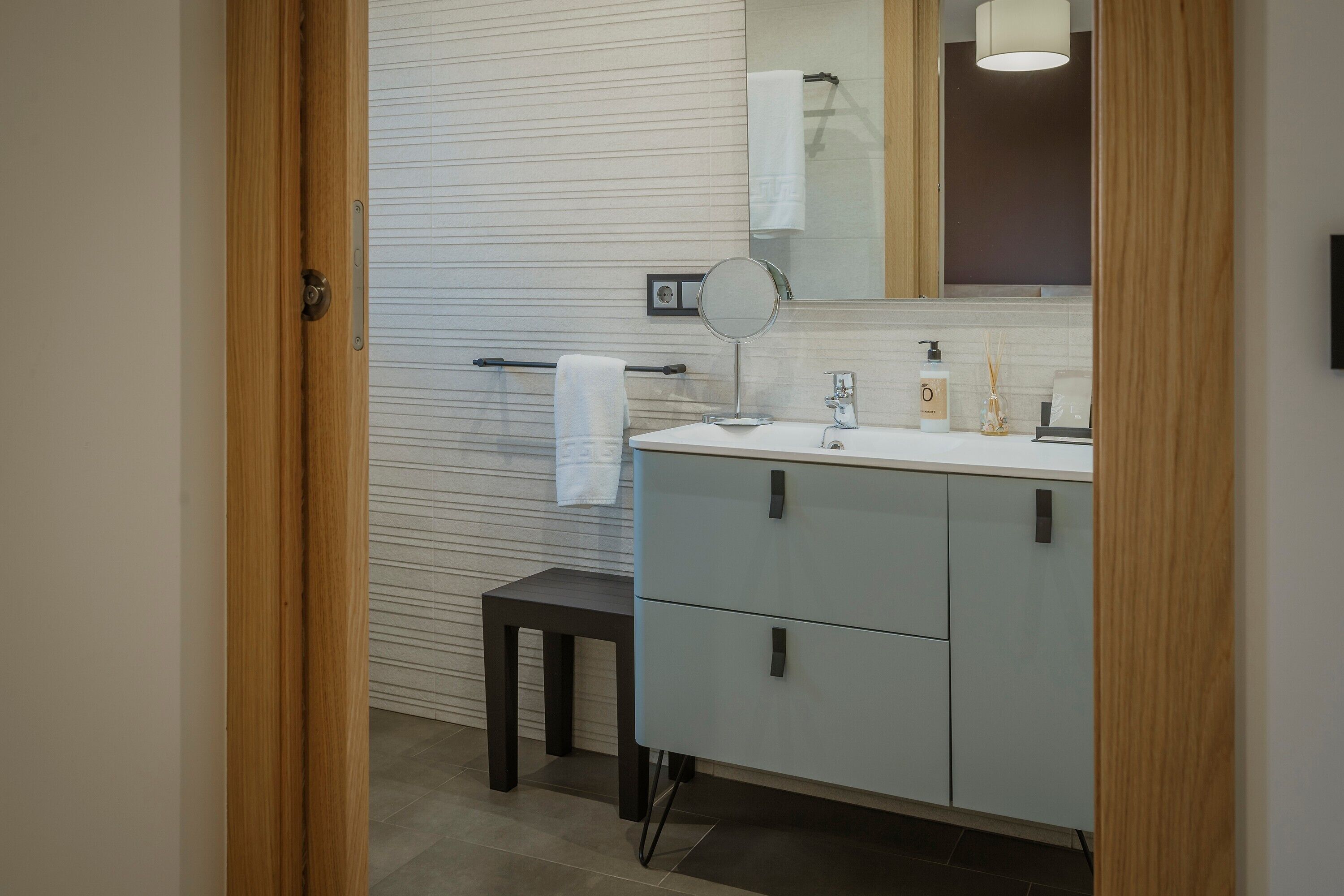 A modern bathroom with a wooden door, a white towel on a towel rack, a round mirror, and a vanity with cabinets.