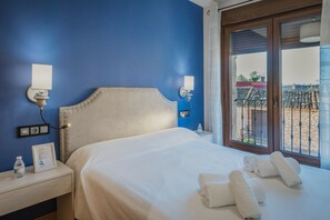 A neatly made bed with white linens and rolled towels, a bedside table with a water bottle and a framed certificate, and a window with a view of a building and a balcony. - Country House 'La Olma' with Private Terrace, Balcony and Wi-Fi (Campillo de Aranda)