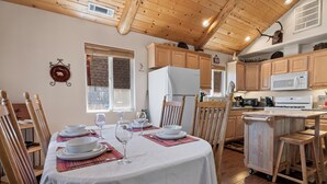Zona pranzo con tavolo imbandito per quattro persone, sedie in legno, tovaglia bianca e angolo cottura con pensili e elettrodomestici in legno.