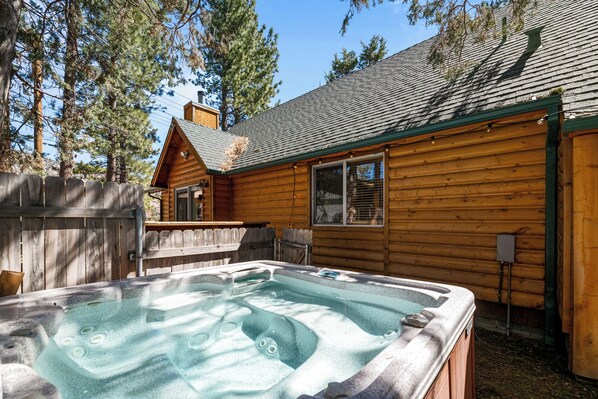 A wooden cabin with a hot tub, surrounded by a wooden fence and tall trees.