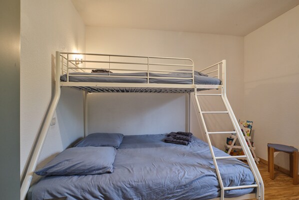 Una cama litera con escalera, un taburete de madera pequeño y un estante con libros.