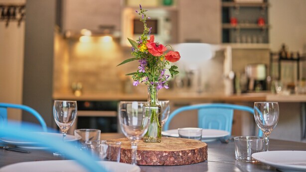 Una mesa de comedor con un centro de flores, copas de cristal y platos, ubicada en una cocina moderna con sillas azules.