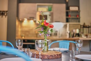 Una mesa de comedor con un centro de flores, copas de cristal y platos, ubicada en una cocina moderna con sillas azules.