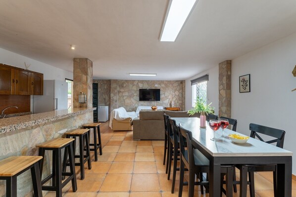 A modern kitchen with a stone backsplash, wooden cabinets, a dining area with a table and chairs, and a TV mounted on the wall.
