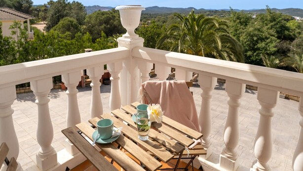 Un balcón con una mesa lista para dos personas, con vistas a un paisaje pintoresco con árboles y colinas.