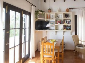 Una habitación con un ventanal grande, televisión, estantería, mesa de comedor con sillas y una planta.