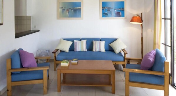 Ein Wohnzimmer mit einem blauen Sofa, zwei Holzsesseln, einem Couchtisch aus Holz und einer Stehlampe.