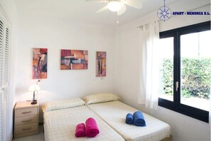 Ein Schlafzimmer mit zwei Betten, einem Deckenventilator und einem Fenster mit Blick auf Grünflächen.