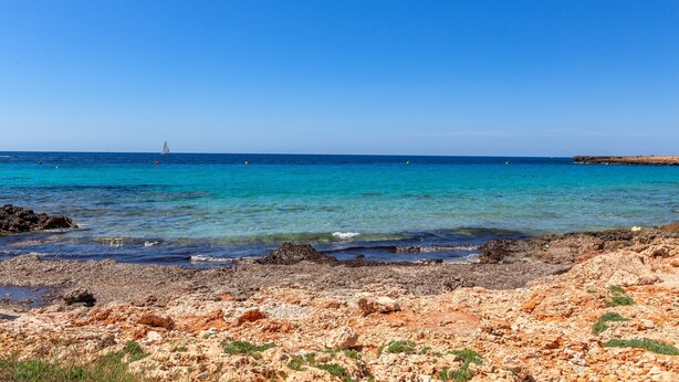 Uma praia rochosa com águas azul-claras e um veleiro ao longe.