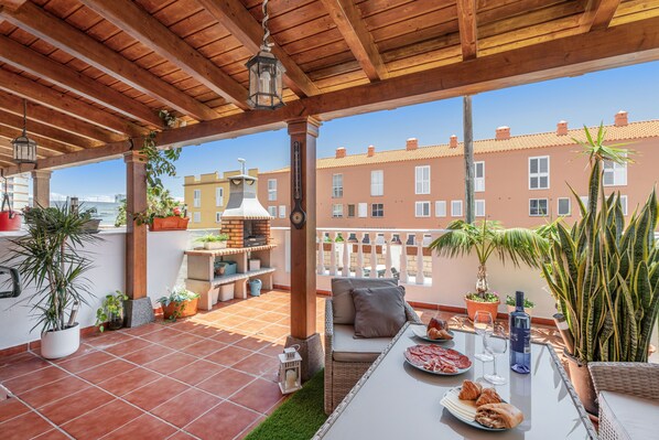 Une terrasse couverte avec une table dressée pour les repas, des sièges d’extérieur, des plantes en pot et une vue sur les bâtiments voisins.