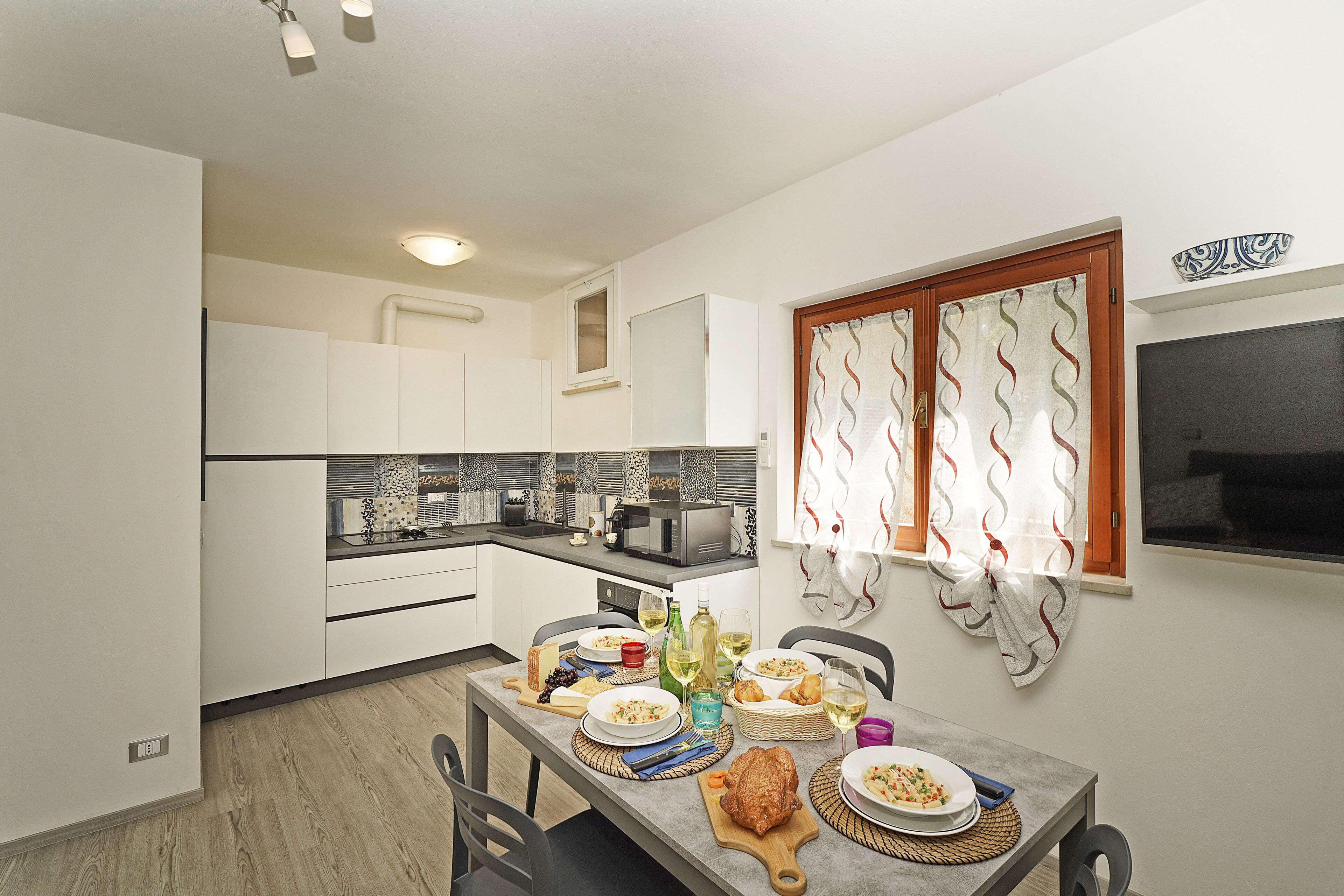 Een kleine keuken met moderne apparatuur, een eettafel gedekt voor een maaltijd, en een raam met decoratieve gordijnen.