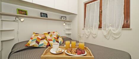 A neatly made bed with a tray of breakfast including toast, fruit, and juice, a bedside table with a lamp, and a window with sheer curtains.
