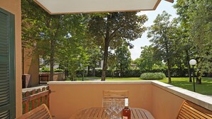 Une table en bois, une bouteille de vin, des verres et une assiette de nourriture sont disposés sur un balcon offrant une vue sur un jardin.