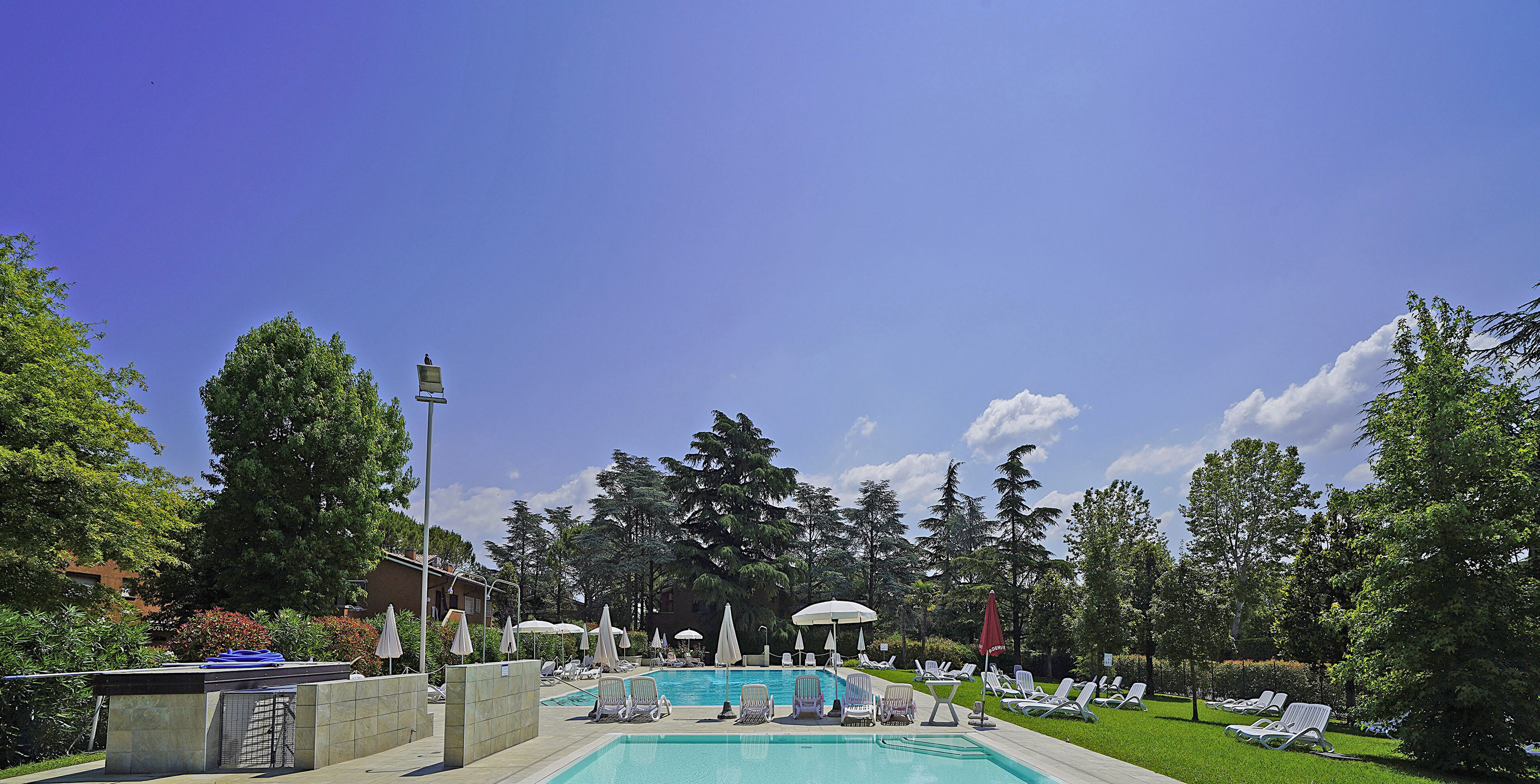 Zona de piscina con sillas de descanso y sombrillas, rodeada de árboles y cielo despejado.