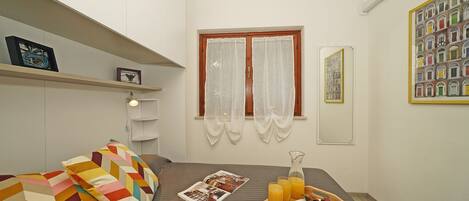 Une petite chambre à coucher, bien rangée, avec un lit, une table de chevet sur laquelle se trouve un plateau de petit-déjeuner, une fenêtre avec des rideaux et une étagère fixée au mur, ornée d’un tableau encadré.