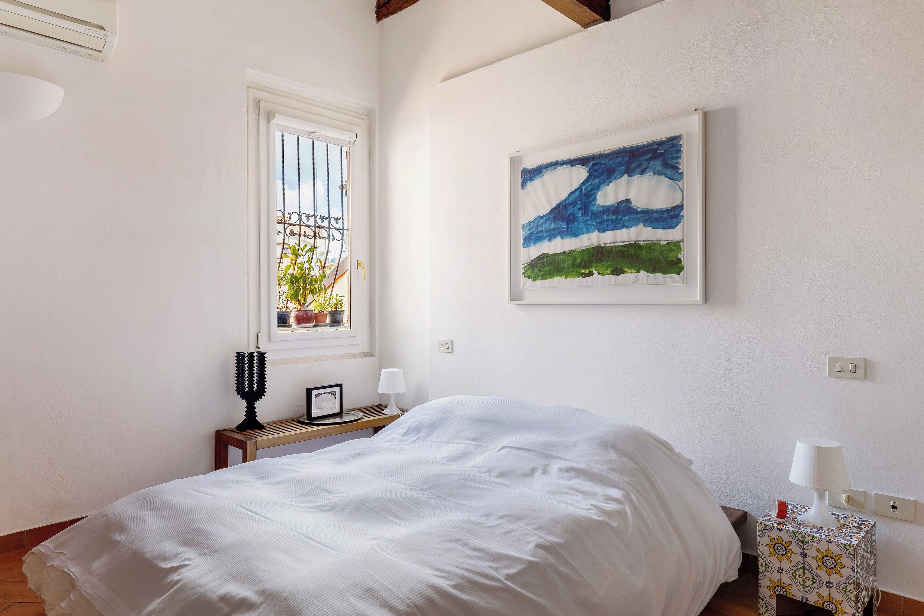 A bedroom with a bed, a window with bars, a bedside table with a lamp, and a painting on the wall.