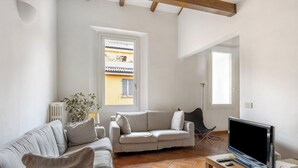 Un salon avec un canapé, un téléviseur et un haut-parleur. Il y a deux fenêtres et un radiateur.