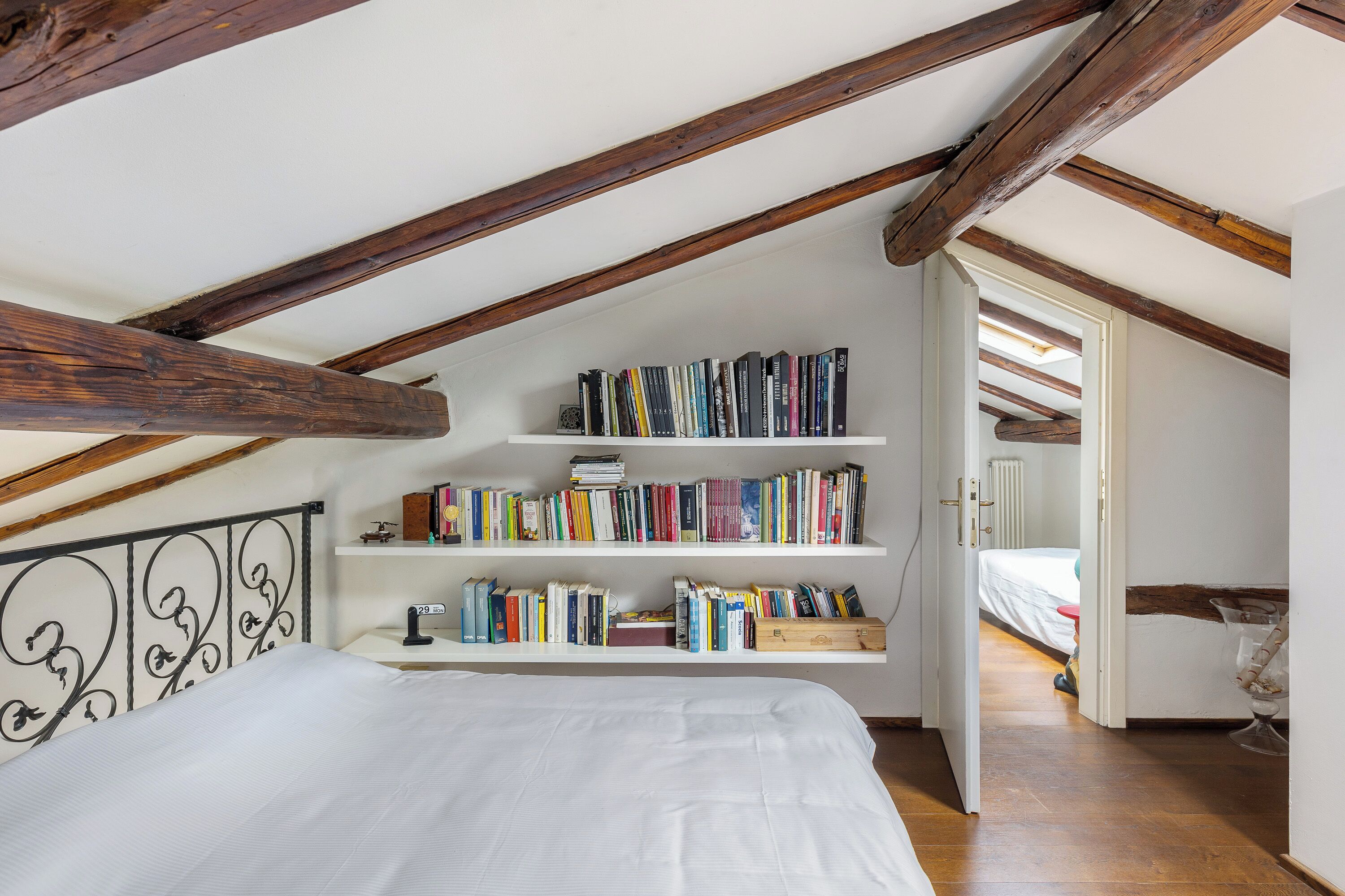 A bedroom with a sloped ceiling, wooden beams, a bed with white bedding, and shelves with books.