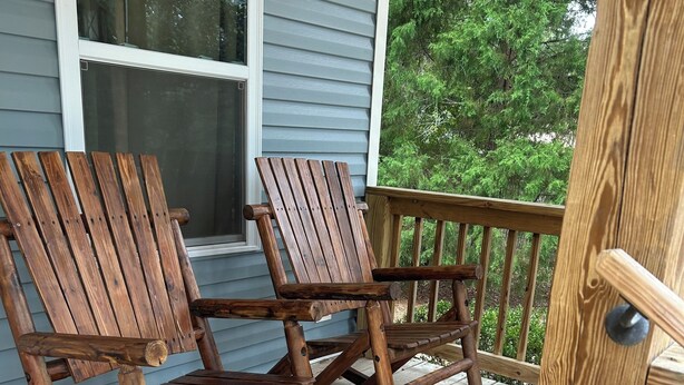 A wooden rocking chair on a porch with a wooden railing and a window in the background.