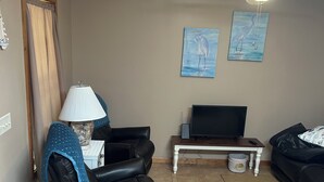A living room with a ceiling fan, a television on a wooden table, a black leather sofa, and two wall-mounted paintings.
