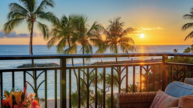 Un balcon agrémenté de meubles en osier, d’un chapeau et d’un vase de fleurs, donnant sur une plage bordée de palmiers et sur l’océan, au moment du coucher du soleil.