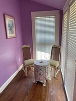 A room with purple walls, a window with blinds, two wicker chairs, and a small round table with a floral tablecloth.