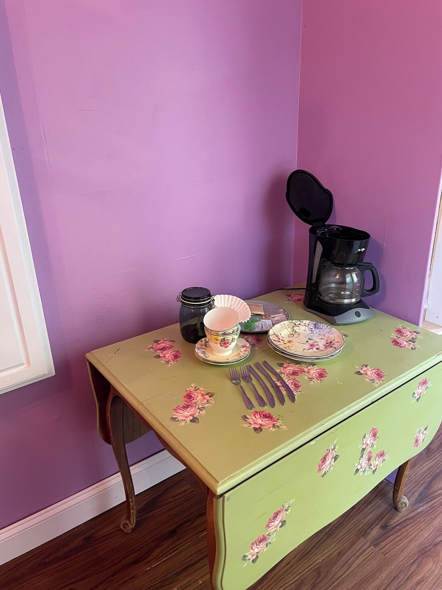 Una mesa de comedor con mantel floral, una cafetera y diferentes platos y utensilios.