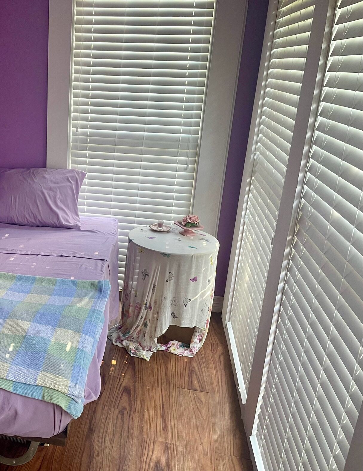 A bedroom with a bed, a small round table with a floral tablecloth, and a window with blinds.