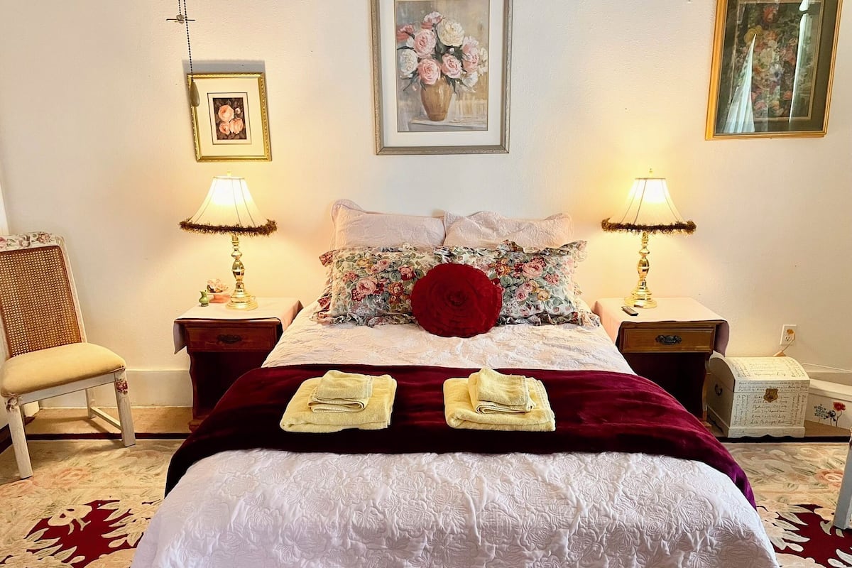 A neatly made bed with a red pillow, two bedside lamps, a chair, and a framed picture on the wall.