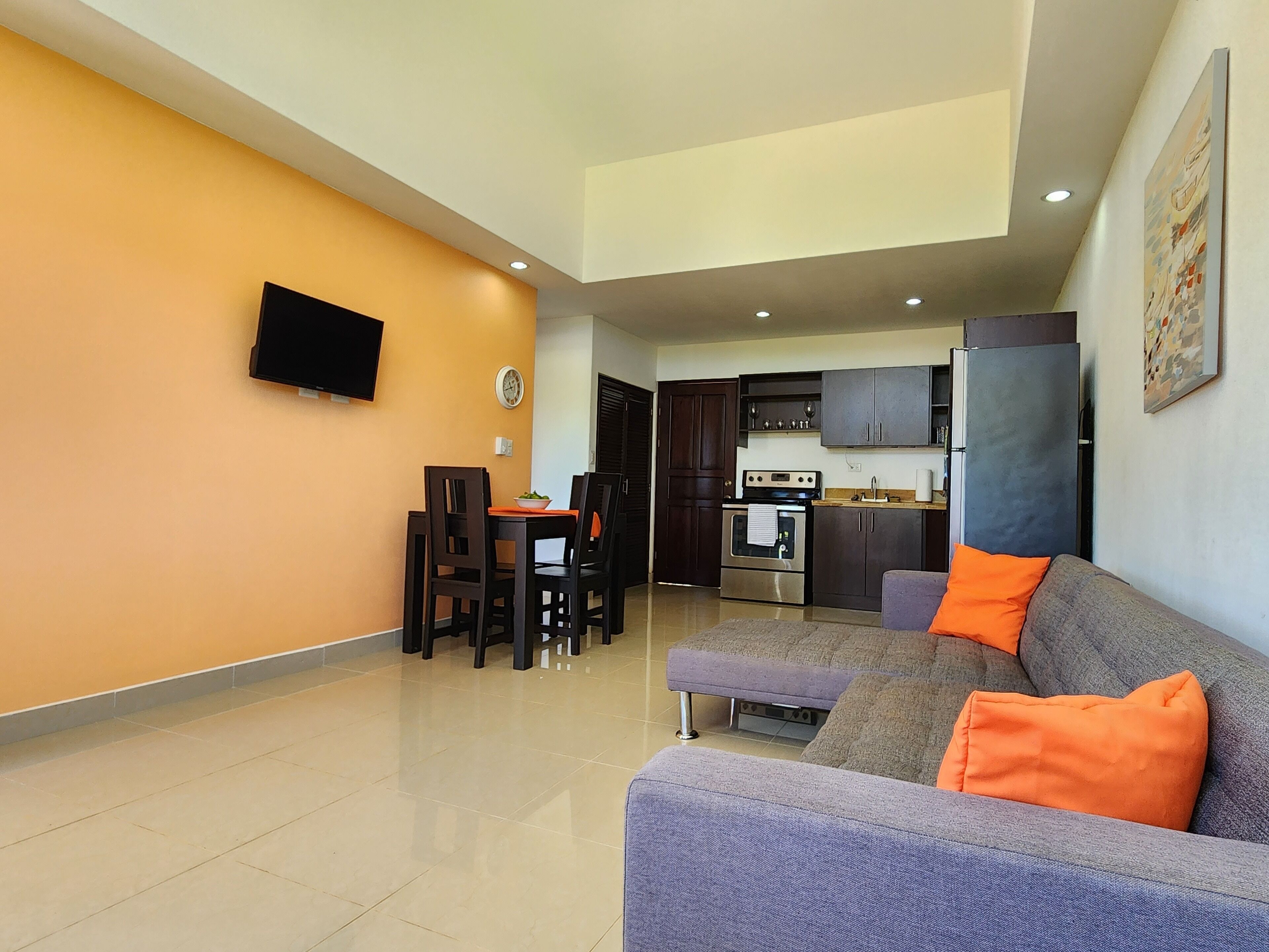 A modern living room with a sofa, orange cushions, a dining area with chairs, a kitchen with cabinets and appliances, and a flat-screen TV mounted on the wall.