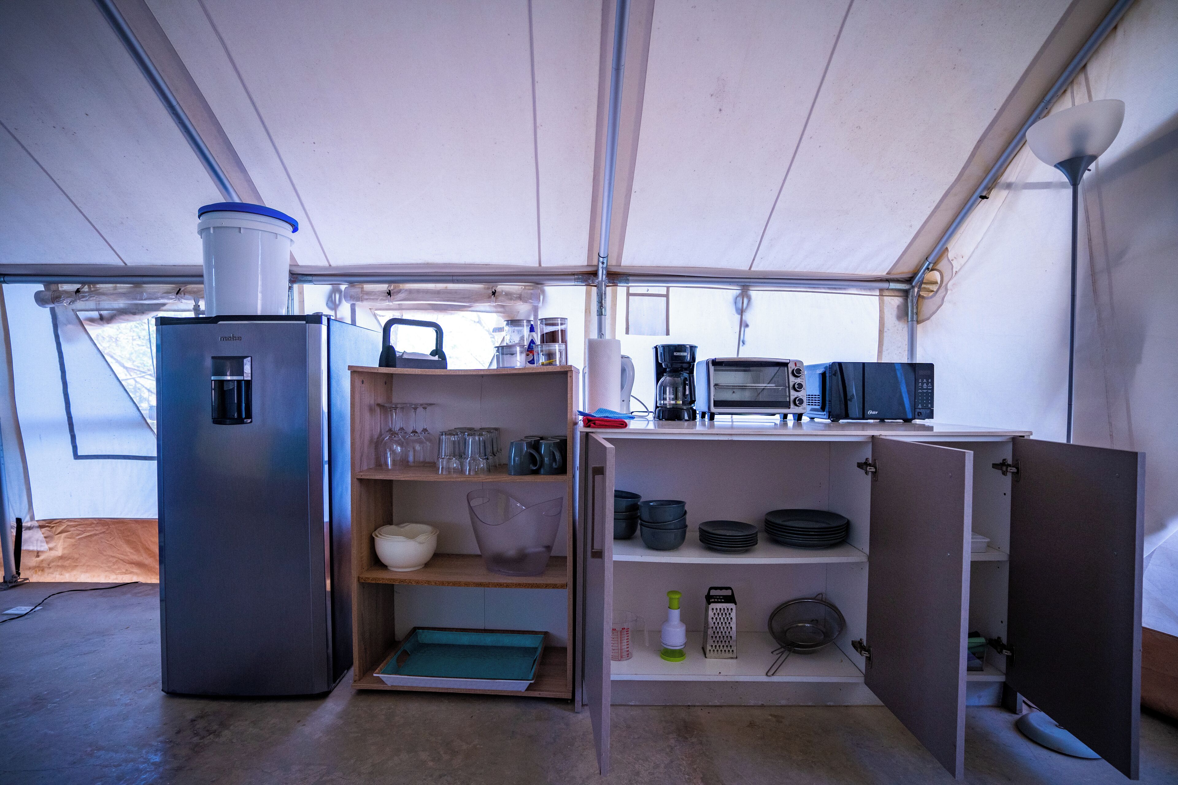 A compact kitchen with a refrigerator, coffee maker, toaster, and shelves with dishes and utensils.