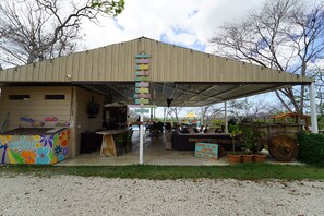A covered outdoor seating area with a rustic metal roof, a colorful recycling bin, and potted plants. - Guanaglamp (Tamarindo)