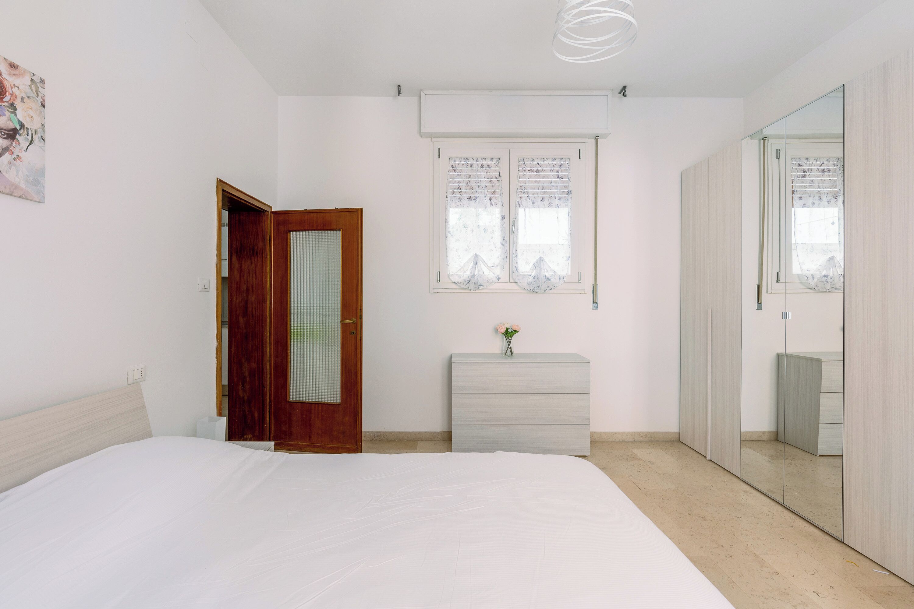 A bedroom with a bed, a wooden door, a dresser, a window with curtains, and a mirror cabinet.