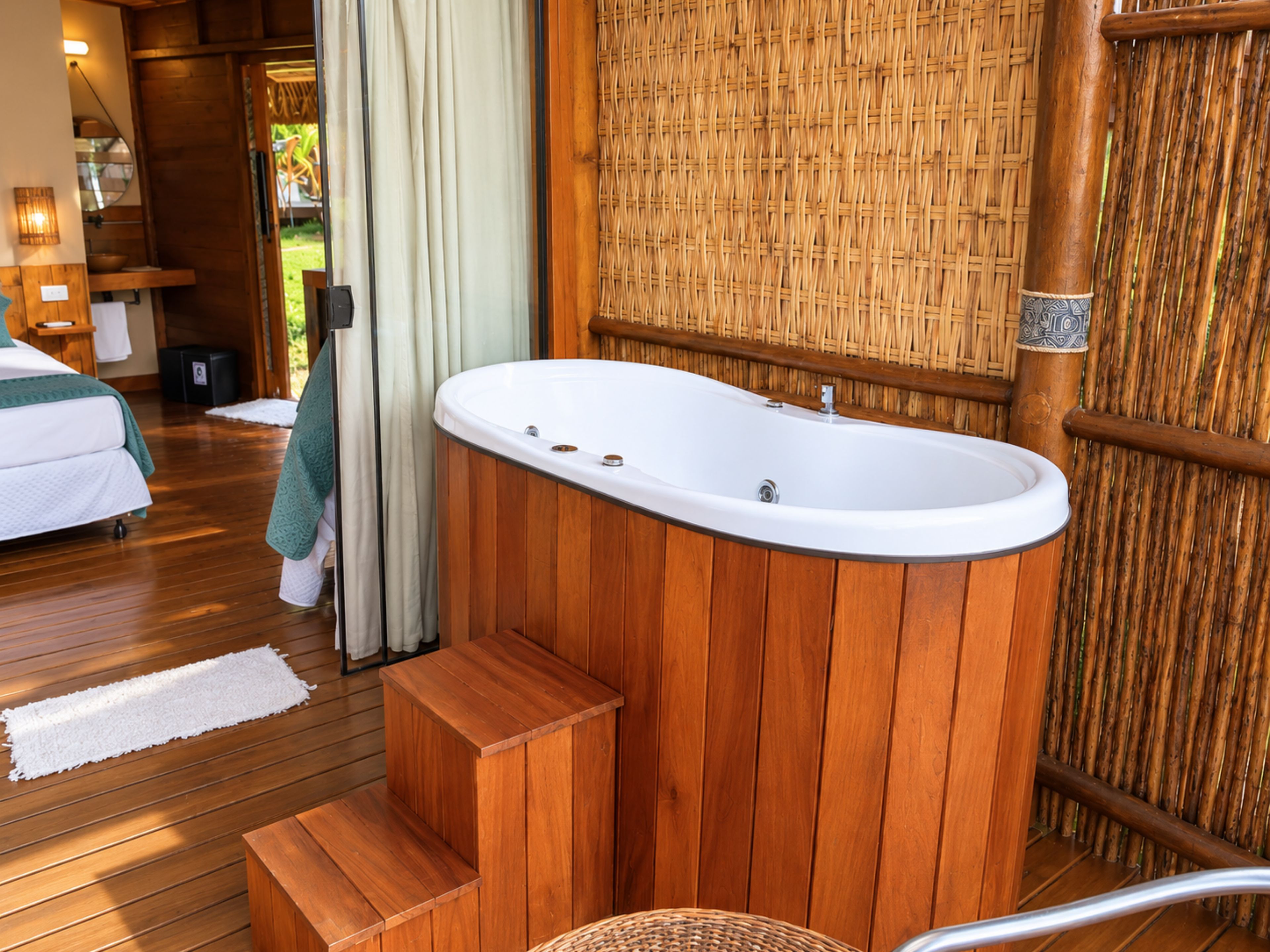 A wooden-floored room with a freestanding bathtub, a bed with a green towel, and a wicker chair.