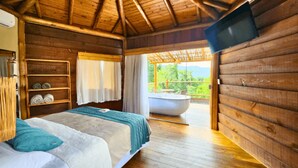 A wooden cabin room with a bed, a shelf with towels, a television, and a view of a balcony with a hot tub. - Tahaa Pousada Boutique (Ilhabela)