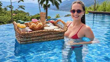 Una piscina con una cesta de mimbre que contiene comida y bebidas, rodeada por un paisaje tropical con montañas al fondo.