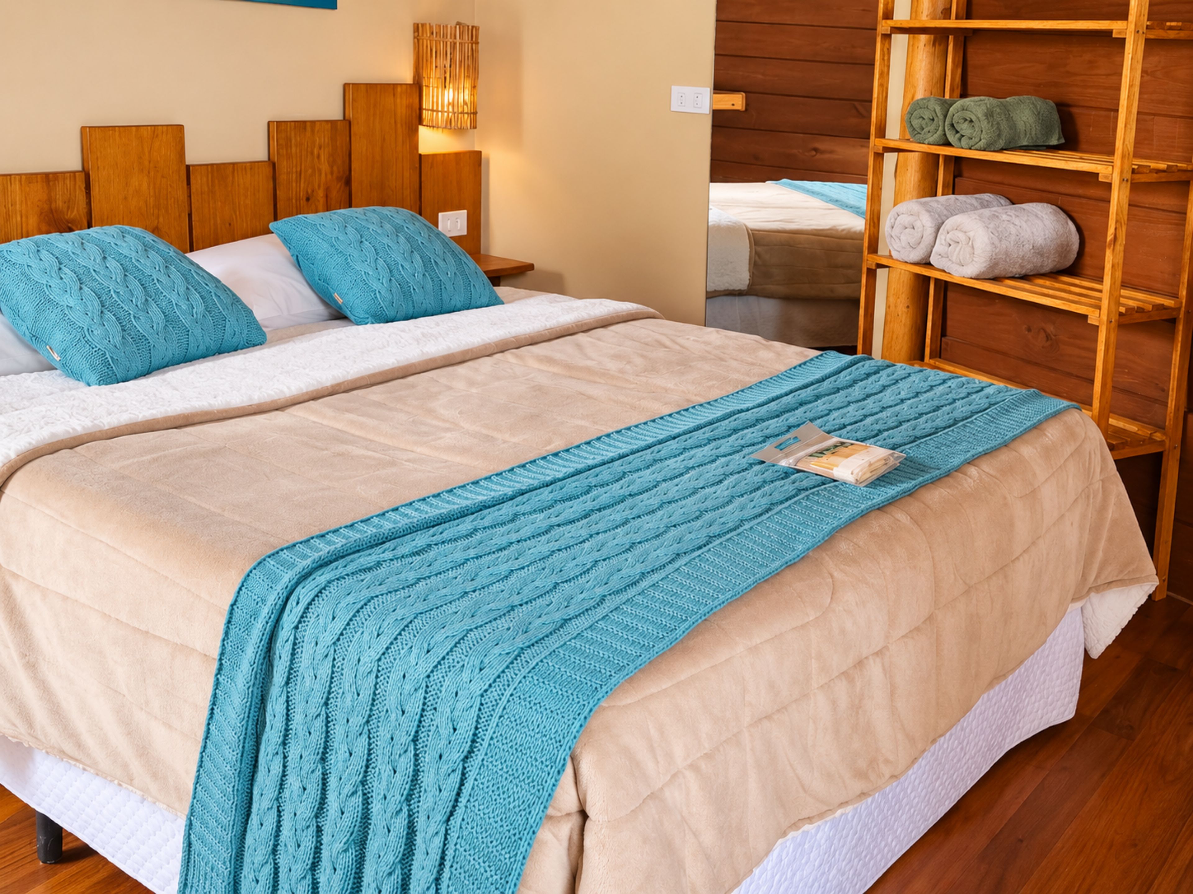 Cama com colcha bege e manta tricotada verde-água, cabeceira de madeira, abajures e prateleira de madeira com toalhas.