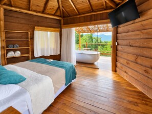 A wooden cabin room with a bed, a shelf with towels, a bathtub, and a TV mounted on the wall. - Tahaa Pousada Boutique (Ilhabela)