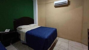 Une petite chambre avec un lit simple, un mur vert, un mur beige et un sol carrelé. On y trouve une table de chevet avec une lampe, une couverture bleue et un appareil de climatisation fixé au mur beige.
