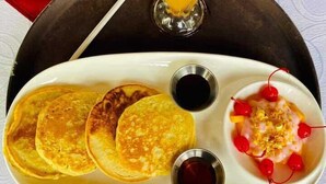 Un plateau de petit-déjeuner avec des crêpes, un verre de jus d’orange et un dessert aux cerises et au sirop.